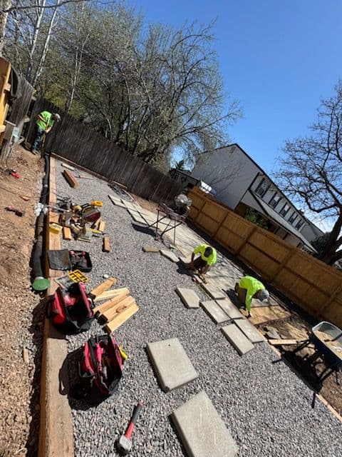 Construction workers laying stones in a backyard landscape renovation under a clear blue sky.