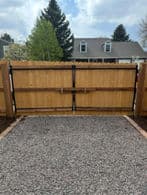 Wooden fence gate with metal hinges, gravel path in front, suburban house in the background.