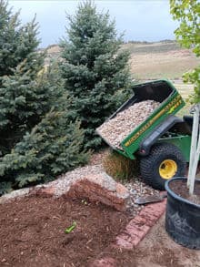 Garden landscaping scene with a cart unloading decorative gravel near evergreen trees.