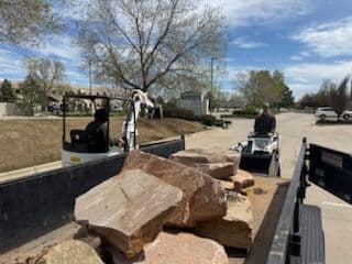 Construction workers operating small machinery to move large rocks at a job site.
