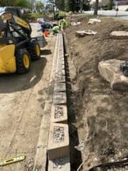 Construction workers laying a stone retaining wall with a skid steer loader nearby.