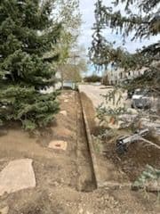 Dirt trench along a landscaped area with trees and pavement in the background.