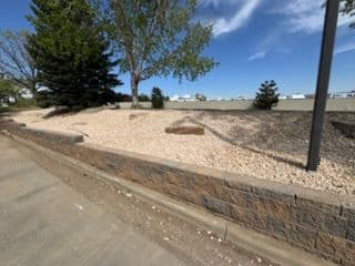 Landscaped area with gravel, trees, and a stone wall under a clear blue sky.